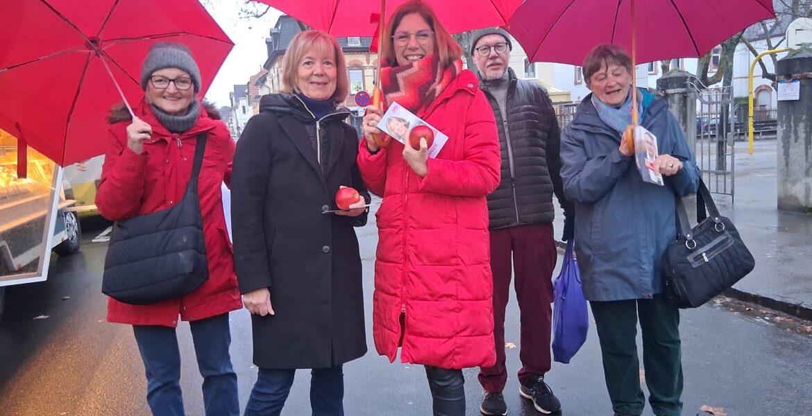 5 Personen stehen auf einer Straße, teilweise mit roten Regenschirmen. Alle schauen in die Kamera.