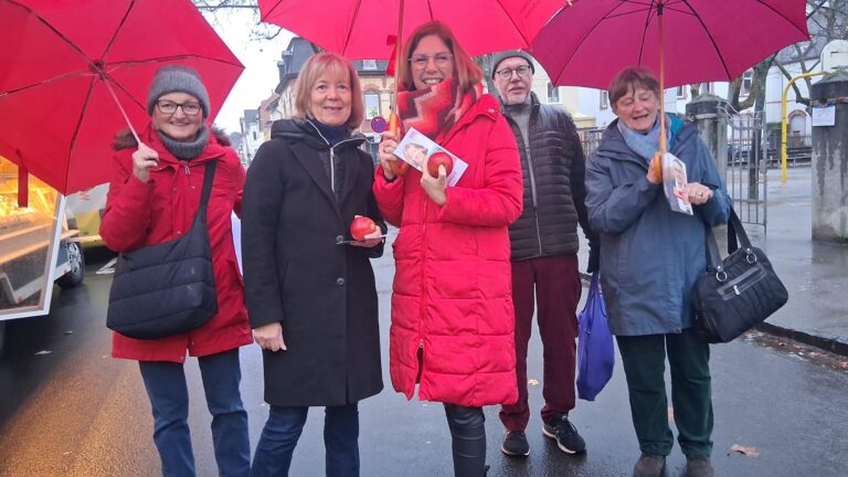 5 Personen stehen auf einer Straße, teilweise mit roten Regenschirmen. Alle schauen in die Kamera.
