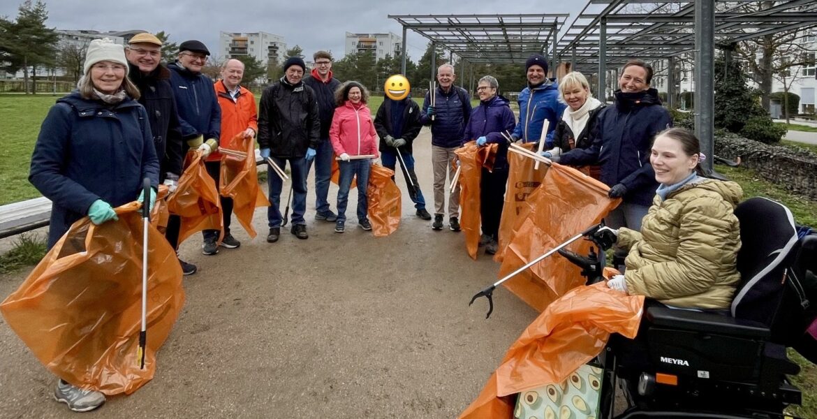 Eine Gruppe von Menschen steht im Halbkreis auf einem Platz. Sie halten orangefarbene Mülltüten und Greifer in den Händen.