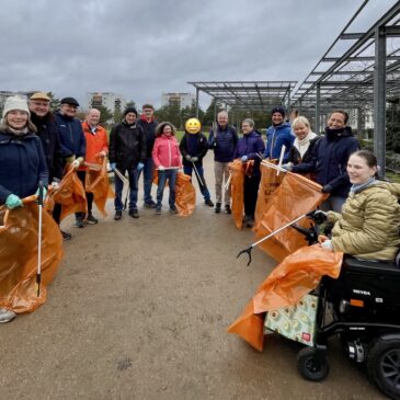 Eine Gruppe von Menschen steht im Halbkreis auf einem Platz. Sie halten orangefarbene Mülltüten und Greifer in den Händen.